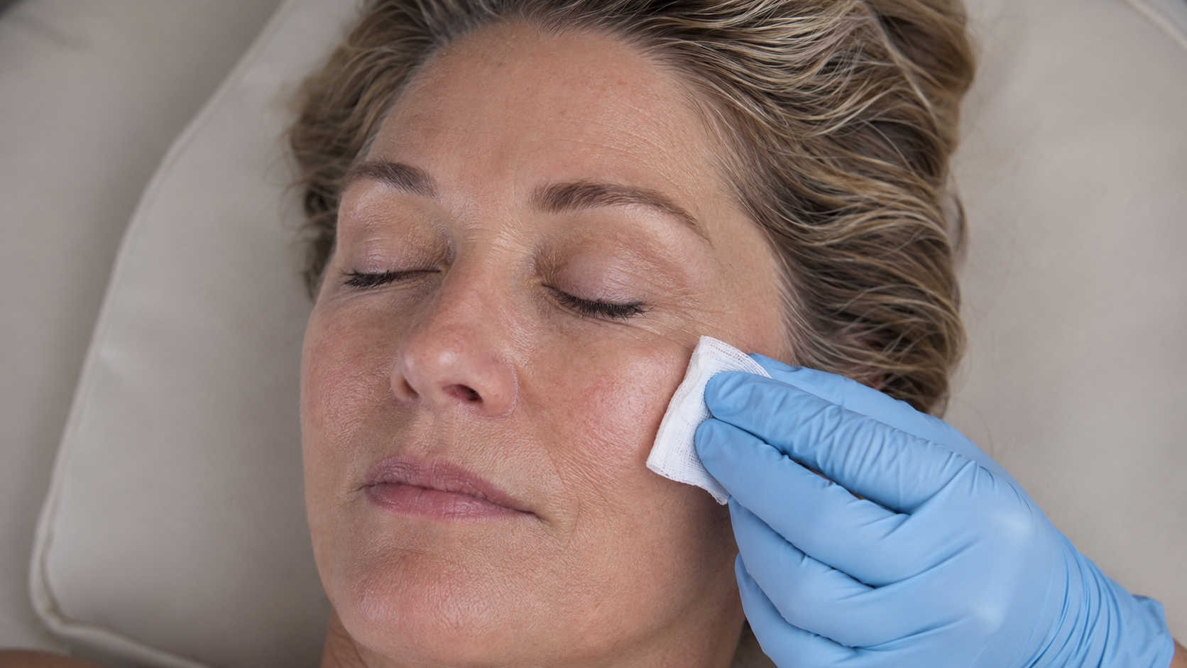 A reclining woman with her eyes closed while a gloved hand gently prepares her cheek with gauze